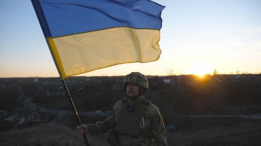 Ukrayna bayrağı sallayan Ukrayna ordusunun askeri. Askeri üniformalı ve kasklı bir adam gün batımında bayrak kaldırdı. Rus saldırısına karşı zafer. İstila direnci konsepti. Yavaş çekim