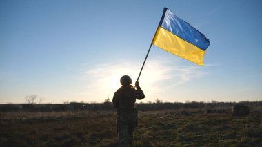 Female ukrainian army soldier walking to the peak of hill that to raise Ukraine flag. Woman in military uniform holding waving flag as symbol of victory against russian aggression. End of war concept.