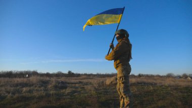Soldier of ukrainian army holding waving flag of Ukraine. Woman in military uniform and helmet lifted up yellow-blue flag. Victory against russian aggression. Invasion resistance concept. Slow motion.