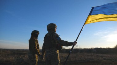 Ukrainian army soldiers stands outdoor at sunset time and waves flag of Ukraine. People in military uniform lifted up flag against blue sky. Victory at war. Resistance to russian invasion. Slow motion.