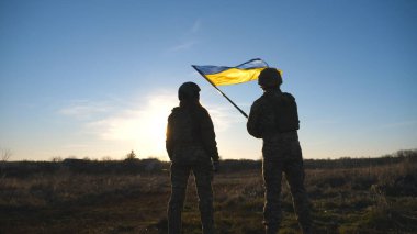 Ukrainian army soldiers stands outdoor at sunset time and waves flag of Ukraine. People in military uniform lifted up flag against blue sky. Victory at war. Resistance to russian invasion. Slow motion.