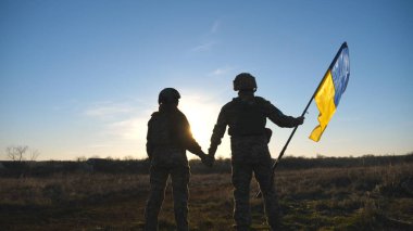 Ukrainian army soldiers stands outdoor at sunset time and waves flag of Ukraine. People in military uniform lifted up flag against blue sky. Victory at war. Resistance to russian invasion. Slow motion.