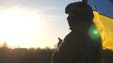 Female ukrainian army soldier using smartphone to read news or messaging with family. Girl in military uniform and helmet holding waving flag of Ukraine. Victory against russian aggression. Close up.