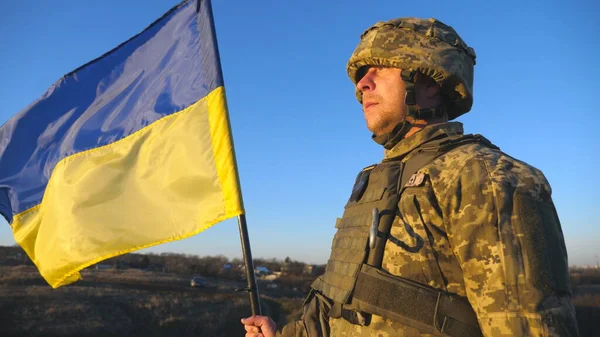 Ukrainian army soldier holding waving flag of Ukraine. Portrait of man in military uniform and helmet lifted up flag in hill. Victory against russian aggression. Invasion resistance concept. Slow mo.