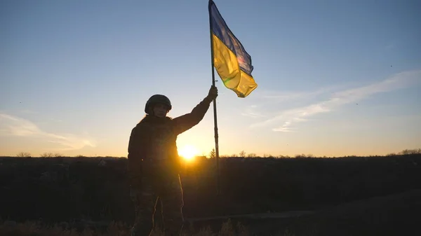 Askeri üniformalı kadın gün batımına karşı duruyor ve Ukrayna bayrağını kaldırıyor. Bayrak sallayan Ukraynalı kadın asker. Rus saldırısına karşı zafer. İstila direnişi kavramı.