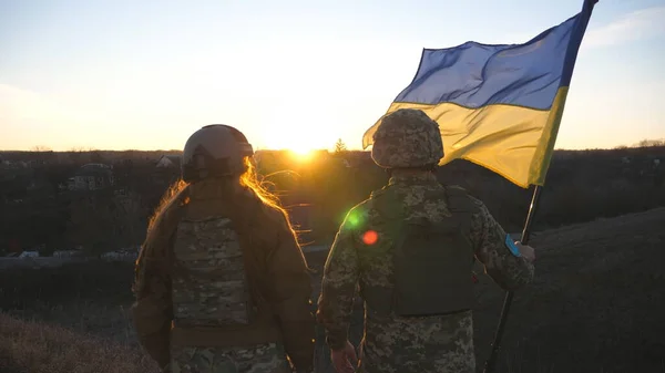 Ukrayna ordusunun askerleri günbatımının arka planına karşı Ukrayna bayrağı çekiyorlar. Askeri üniformalı insanlar sarı-mavi bayrak kaldırdı. Rus saldırısına karşı zafer. İstila direnişi kavramı.