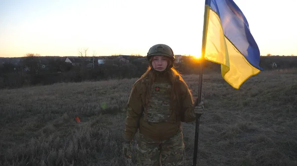 Female military medic walking at the field with waving flag of Ukraine. Ukrainian girl in uniform and helmet going with lifted up flag at sunset time. Victory against russian aggression. Slow motion.