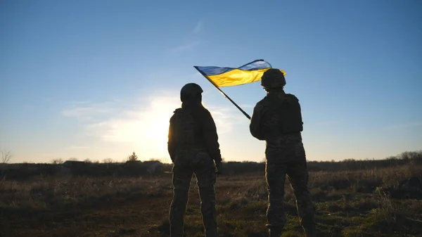Ukrainian army soldiers stands outdoor at sunset time and waves flag of Ukraine. People in military uniform lifted up flag against blue sky. Victory at war. Resistance to russian invasion. Slow motion.