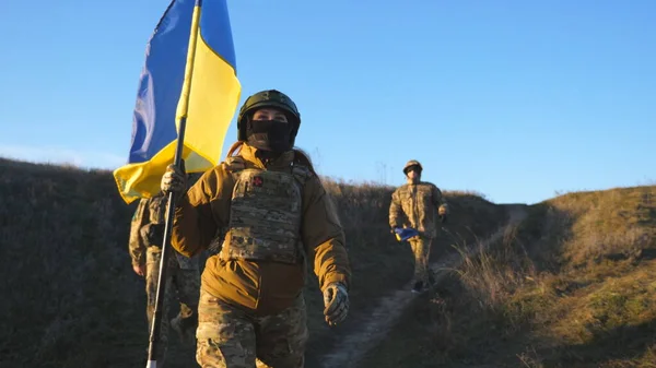 Soldiers of ukrainian army walks at field with flag of Ukraine. People in military uniform waving national yellow-blue flag as sign of victory against russian aggression. Invasion resistance concept.