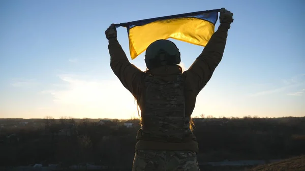 Ukrainian military medic holding waving flag of Ukraine. Female army soldier in uniform and helmet lifted up flag at the hill. Victory against Russian aggression. Invasion resistance concept. Slow mo.