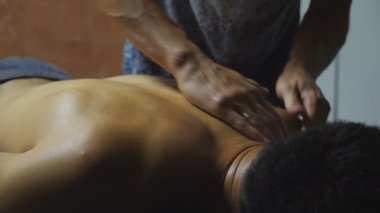 Male arms of masseur massaging shoulders of muscular sportsman in salon. Hands of professional massagist doing healing rubdown of back to young athlete in parlor. Concept of skincare. Slow motion.