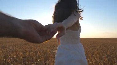 Young woman holding male hand and running on endless wheat field at sunset background. Happy girl looking back into camera while leading her boyfriend along barley meadow. Freedom concept. Slow motion