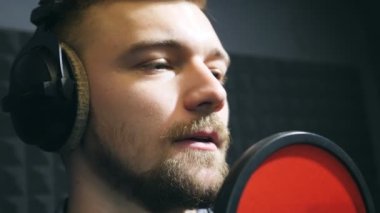 Kulaklıklı erkek şarkıcı ses stüdyosundaki mikrofona şarkı söylüyor. Genç adam duygusal olarak yeni bir melodi ya da şarkı kaydediyor. Yaratıcı bir müzisyenin çalışması. Gösteri dünyası konsepti. Yavaş çekim.