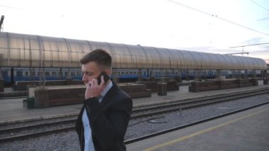 Successful businessman walking through railway station and talking on phone. Handsome man strolling near railway and having work conversation. Guy goes outdoor speaking on cellphone. Slow motion.
