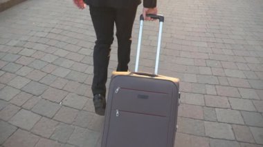 Legs of successful businessman walking on city street and pulling suitcase on wheels at sunset. Young successful man going with his luggage on urban environment. Travel concept. Back view Slow motion.