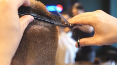 Male hands of hairdresser combing and cutting hair of customer with hairbrush and scissors in salon. Arms of barber trimming hair of his client in barbershop. Hairstyling process. Close up Slow motion