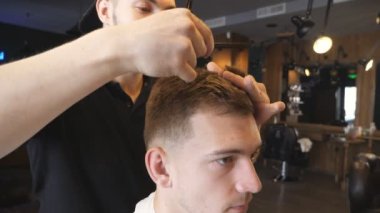 Close up of male barber combing and cutting hair of his young customer by hairbrush and scissors in barbershop. Handsome guy getting his hair dressed at barber shop. Hairstyling process. Slow motion.