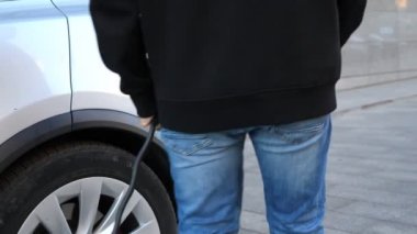 Elektrikli bir arabanın kapağını açıp şarj aletini takan bir adam. Elektrikli bir araca yakıt ikmali işlemi başlatılıyor. Çevre dostu sıfır emisyonlu yeni teknoloji. Kapat.