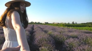 Attractive woman in hat holding male hand and walking through lavender field. Young couple strolling among blooming flower meadow. Boy and girl relaxing at nature. Scenic background. POV Slow motion.