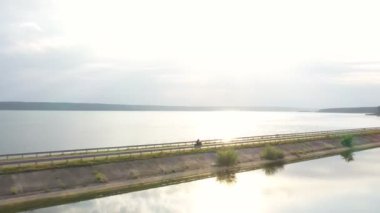 Arka planda güneş ışığı olan baraj güzergahında motosiklet süren motorcuya hava görüntüsü. Motosikletli göl kenarındaki yolda motosikletiyle yarışıyor. Yolculuk sırasında bisiklet süren bir adam. Macera kavramı. Üst görünüm