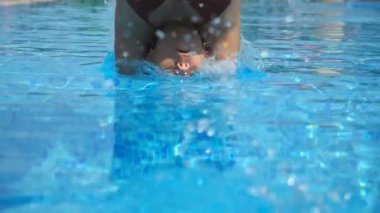 Young attractive woman diving in pool and then emerges from under the water. Girl in swimsuit swimming in basin and enjoying holiday. Lady relaxing during summer vacation. Bottom view Slow mo Close up