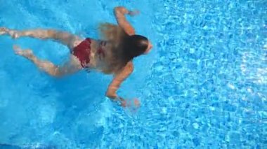 Young woman swimming in pool with crystal clear water at sunny day. Attractive girl relaxing during refreshing in basin. Concept of summer vacation. Slow motion Top view.