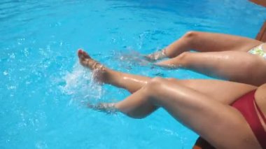 Kadın bacakları otel havuzunda kristal berraklığında su sıçrayan. Havzakenarında oturan ve güneşli bir günde tatillerinin tadını çıkaran iki arkadaş. Yaz tatili veya tatil kavramı. Yavaş çekim Yan görünüm.