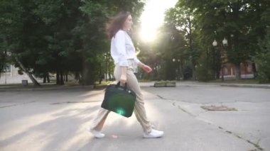 Female office worker with bag going on city street with sun shining at background. Businesswoman in formal wear stepping through urban district. Side view to confident lady walks outdoor. Slow motion.