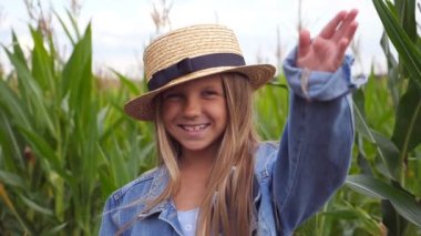 Happy small girl looking into camera and straightening her long blonde hair against the background of corn field. Portrait of joyful kid in straw hat standing in the meadow and laughing. Close up.