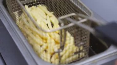 Erkek el aşçı kafesi patates kızartmasıyla sallayarak kızgın yağda hazırlanıyor. Şefin kolu patates ızgarasını fritözün içine indiriyor. Mutfakta fast food hazırlama süreci. Üst görünüm.