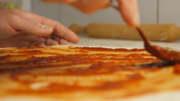 台所のテーブルでスプーンを使用してピザ生地にケチャップを適用調理の男性の腕 料理の表面に金属の形でペストリーにトマトソースを広げるシェフの手 食物の調製の概念 — ストック動画