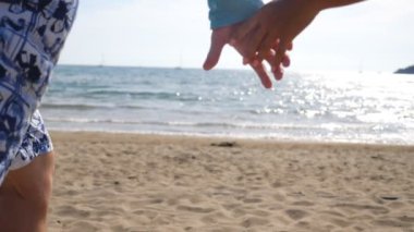 Young couple taking hands of each other and running along beach to sea. Pair rejoicing freedom or their summer vacation at ocean coast. Travelers enjoying new scenic place at seacoast. Slow motion.