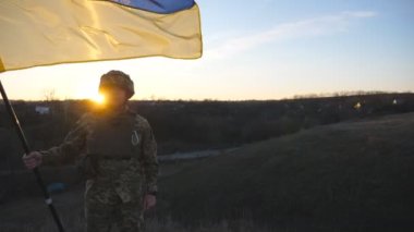 Ukrayna bayrağı sallayan Ukrayna ordusunun askeri. Askeri üniformalı ve kasklı bir adam gün batımında bayrak kaldırdı. Rus saldırısına karşı zafer. İstila direnci konsepti. Yavaş çekim