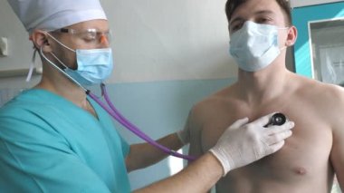 Steteskoplu sıhhiyeci, COVID-19 semptomları arayan hastaların akciğerlerini dinliyor. Koronavirüs zatürresi belirtileri gösteren steteskoplu bir doktor. Doktor, hastanedeki adamın göğsünü kontrol ediyor..