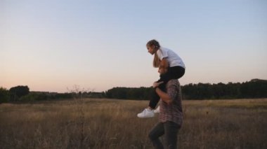 Genç baba, çayırda omuzlarında oturan tatlı küçük kızıyla gidiyor. Mutlu baba ve küçük çocuğu alacakaranlıkta çim tarlasında birlikte vakit geçiriyorlar. Aile mutluluğu konsepti. Dolly vurdu..