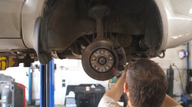 Yetişkin oto tamircisi garajda eski bir arabanın vidalarını söker. Atölyede kaldırma aracının altında çalışan, üniformalı profesyonel sakallı bir tamirci. Otomobil bakımı kavramı. Kapat..