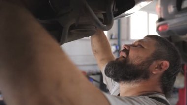 Garajdaki bir kaldırma aracının altında çalışan sakallı araba tamircisinin profili. Üniformalı profesyonel bir tamirci atölyede araba tamir ediyor. Otomobil bakımı kavramı. Yavaşça kapat..