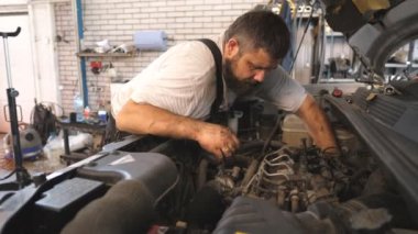 Tamirci atölyede ya da serviste bir arabanın motorunu tamir ediyor. Profesyonel oto tamircisi araç kaputu altında çalışır. Sakallı adam garajda otomobil motorunu kontrol ediyor. Bakım ve tanı kavramı.