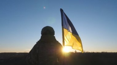 Kamuflaj üniformalı genç bayan asker Ukrayna bayrağını kaldırıyor. Ukraynalı asker kadın gün batımında mavi-sarı bayrak sallayarak tepenin zirvesinde duruyor. Rus istilasına karşı direniş.