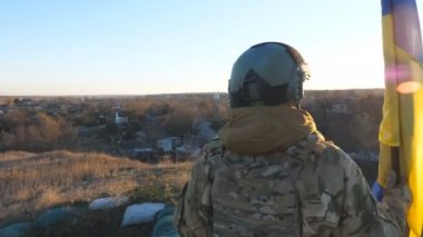 Kamuflaj üniformalı genç bayan asker Ukrayna bayrağını kaldırıyor. Ukraynalı asker kadın gün batımında mavi-sarı bayrak sallayarak tepenin zirvesinde duruyor. Rus istilasına karşı direniş.