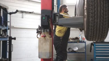 Üniformalı bir tamirci atölyede ya da garajda araba arızalarını tamir ediyor. Araba servisinde kaldırma aracıyla çalışan profesyonel bir tamirci. Düzenli otomobil bakımı kavramı. Yavaş çekim.