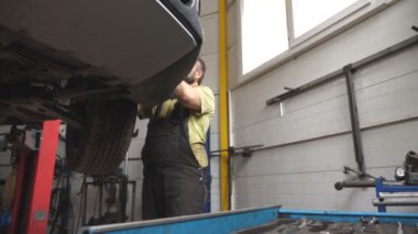 Üniformalı bir tamirci atölyede ya da garajda araba arızalarını tamir ediyor. Araba servisinde kaldırma aracıyla çalışan profesyonel bir tamirci. Düzenli otomobil bakımı kavramı. Yavaş çekim.