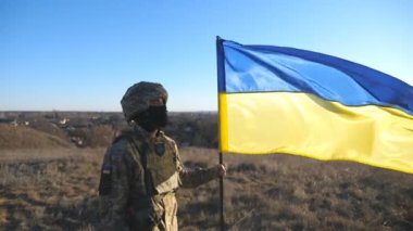 Askeri üniformalı ve miğferinde Ukrayna bayrağı sallayan bir erkek asker resmi. Ukraynalı ordu mensubu ulusal bayrağı kaldırıyor. Rus saldırısına karşı zafer. Yavaş çekim.