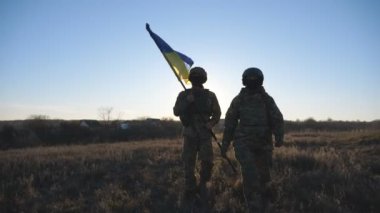 Askeri üniformalı genç kadın ve adam gün batımında çayırda mavi-sarı pankartıyla gidiyorlar. Ukrayna bayrağıyla yürüyen Ukrayna ordusunun kadın ve erkek askerleri. Rus işgaline direniş