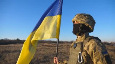 Askeri üniformalı ve kasklı erkek asker Ukrayna bayrağı sallıyor. Ukraynalı ordu mensubu ulusal bayrağı kaldırıyor. Savaşın sonu. Bağımsız ülkenin topraklarına saldırı.