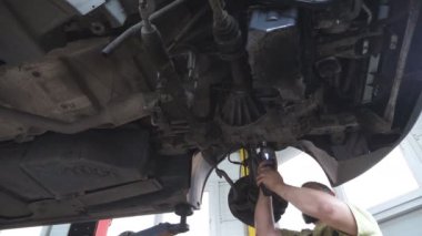 Tamirci araba tamirhanesinde ya da garajda araba tamir ediyor. Atölyede kaldırma aracının altında çalışan profesyonel sakallı oto tamircisi. Düzenli otomobil bakımı kavramı. Yavaş çekim.
