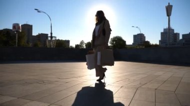 Fashionable lady carries paper packets walking along city square. Young beautiful woman holds shopping bags going at urban street after purchases. Concept of leisure, sales and discounts. Slow motion.