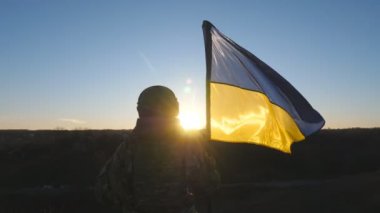 Ukraynalı asker kadın gün batımında mavi-sarı bayrak sallayarak tepenin zirvesinde duruyor. Kamuflaj üniformalı genç bayan asker Ukrayna bayrağını kaldırıyor. Rus istilasına karşı direniş.