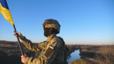 Askeri üniformalı erkek askerin Dolly çekimi Ukrayna bayrağını sallayarak tepenin zirvesinde duruyor. Ukraynalı asker mavi gökyüzüne mavi-sarı pankart kaldırdı. Rus istilasına karşı direniş.