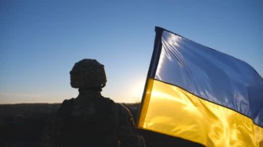 Askeri üniformalı genç Ukraynalı asker gün batımına karşı mavi-sarı pankart kaldırıyor. Erkek asker tepenin zirvesinde Ukrayna bayrağını sallayarak Rus saldırısına karşı zaferin şerefine duruyor..